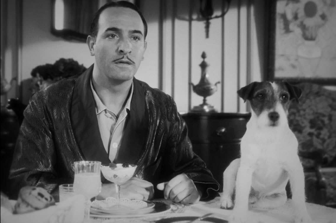 Hombre sentado en una mesa con una mirada expresiva y un cachorro a su lado con cara de sorpresa.