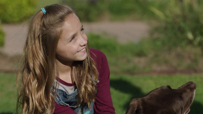 Menina loira olhando para o lado e sorrindo, com um cachorro castanho