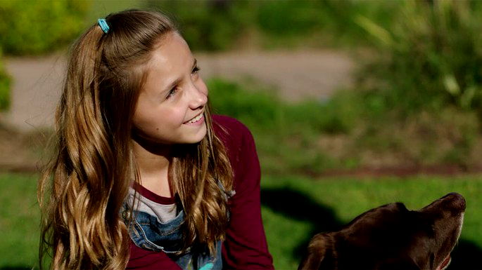 Menina loira olhando para o lado e sorrindo, com um cachorro castanho