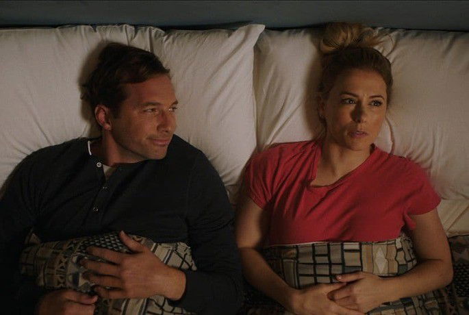 Casal deitado na cama; ela com expressão preocupada e ele olhando para ela.