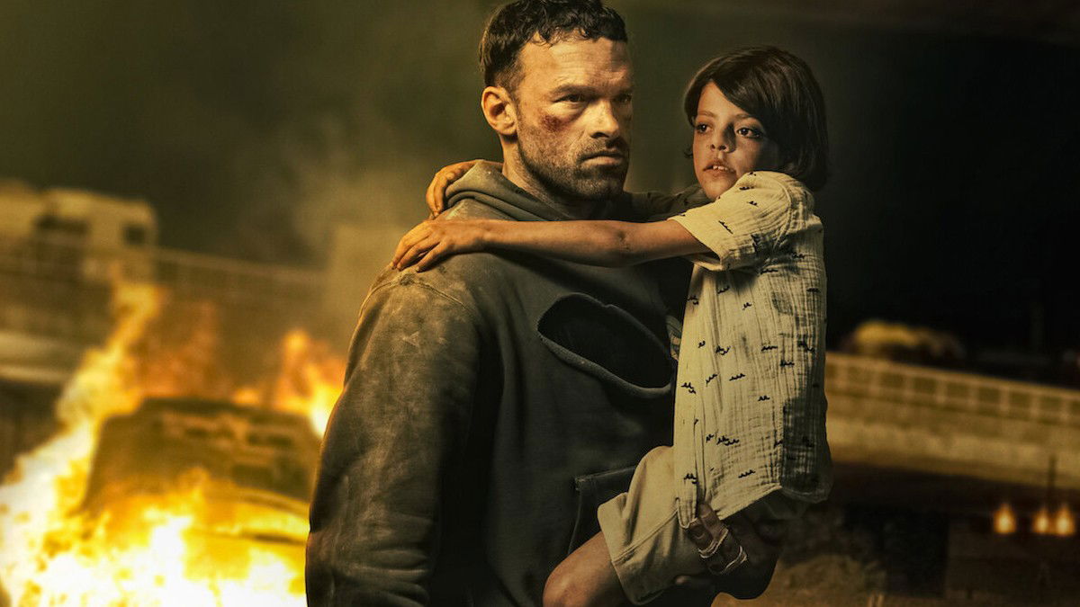 cena do filme agente infiltrado mostra um homem segurando no colo um  menino em meio a um cenário de guerra