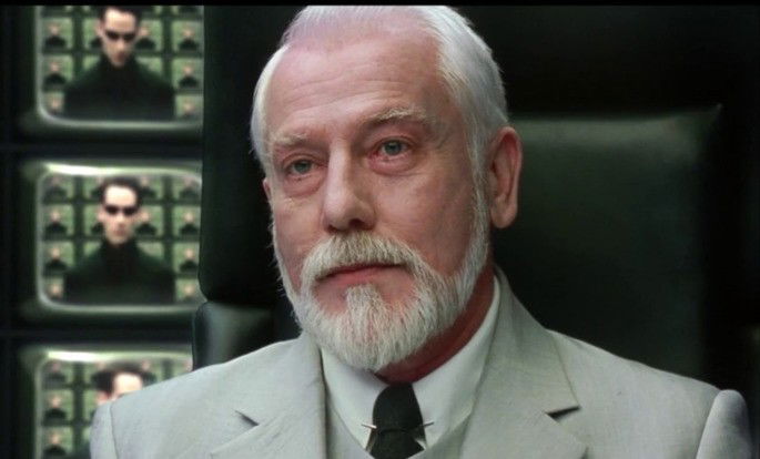 Homem de cabelo e barba branca, com várias televisões atrás.