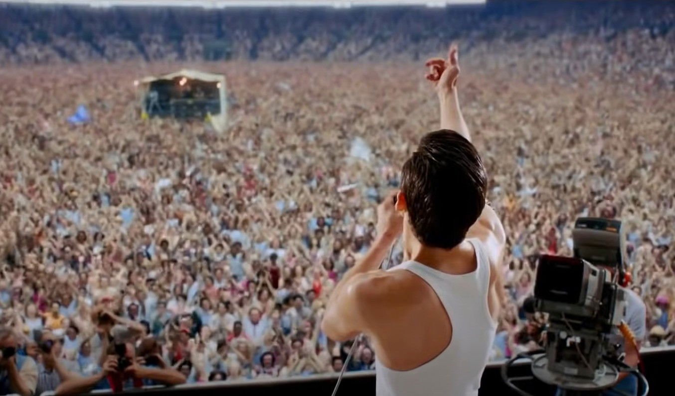 Show do Queen no Live Aid no filme. Ator caracterizado como Freddie Mercury de costas e plateia ao fundo