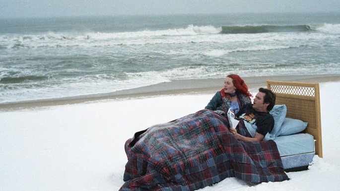 Casal deitado numa cama, no meio da praia.