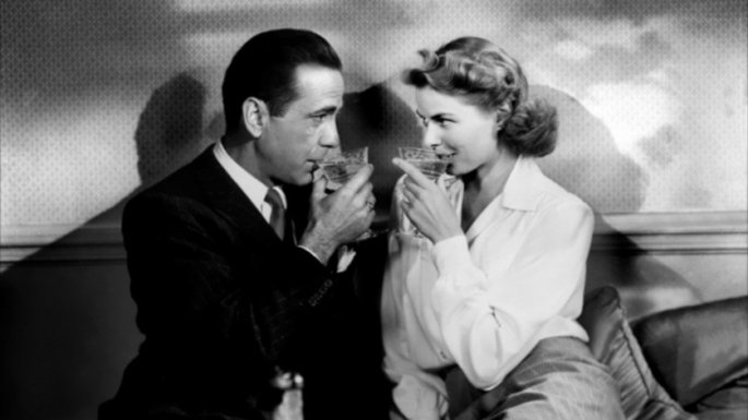 Casal de protagonistas bebendo um drink e se olhando nos olhos.