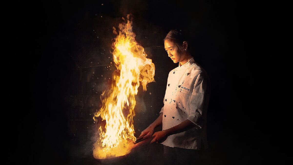 cena do filme Fome de sucesso mostra mulher oriental com roupa de cozinheira em frente uma fogueira