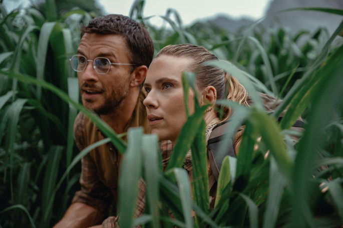 Mulher e homem escondidos no mato, observando os dinossauros.