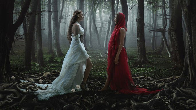 Duas mulheres, frente a frente, numa floresta. Uma usa um vestido vermelho e a outra um vestido branco.