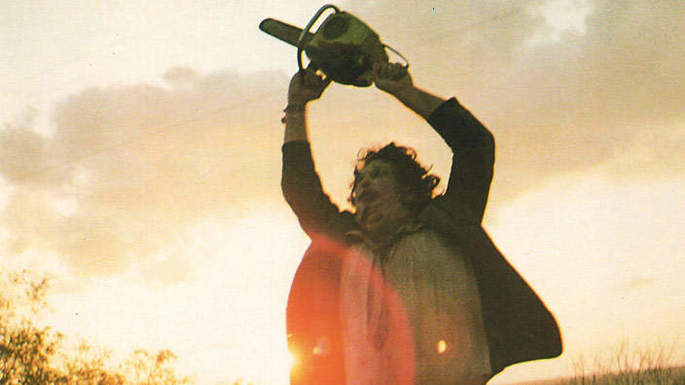 Homem numa estrada, com máscara assustadora, levantando uma serra elétrica.