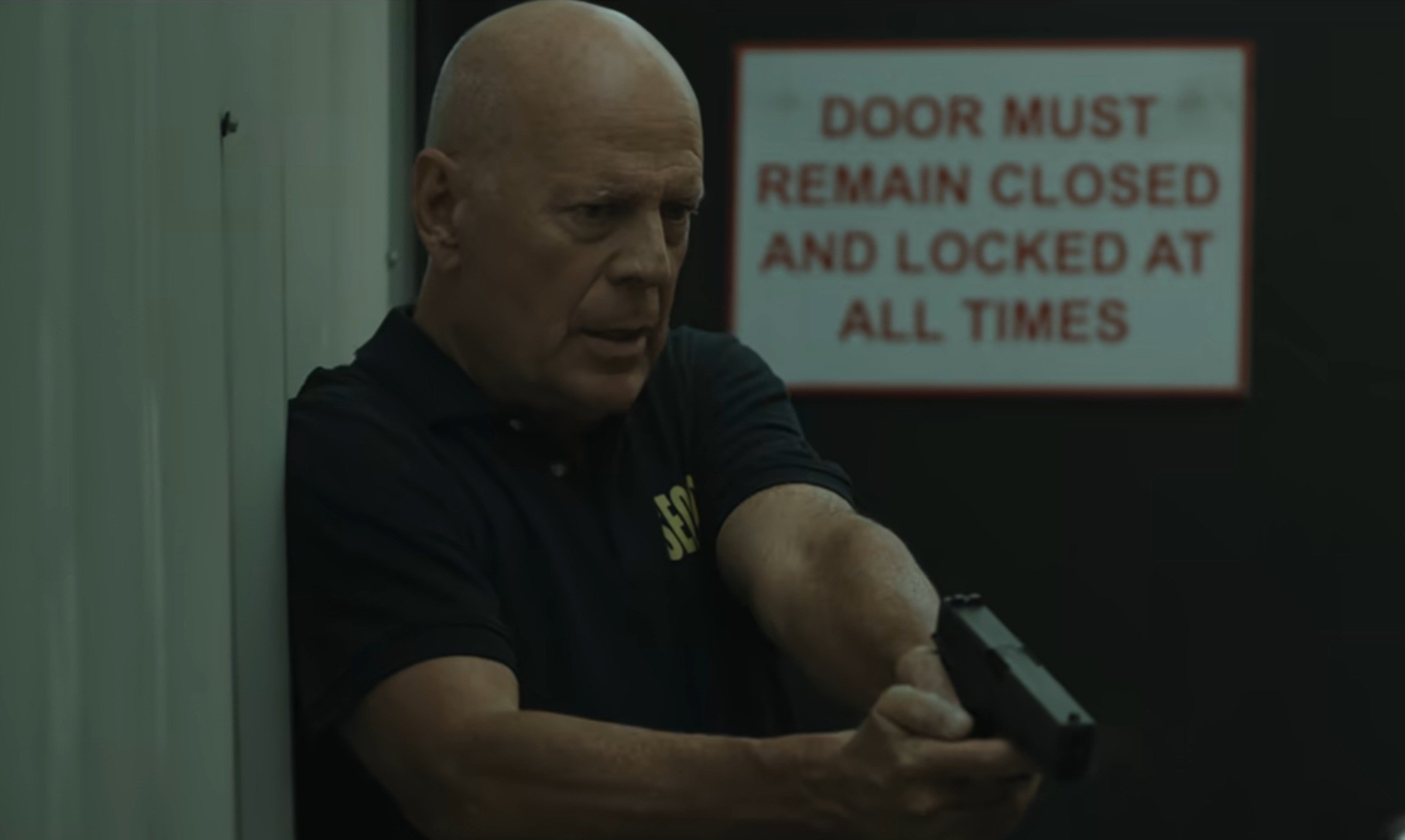 Bruce Willis com arma em punho e roupa de segurança, aponta arma