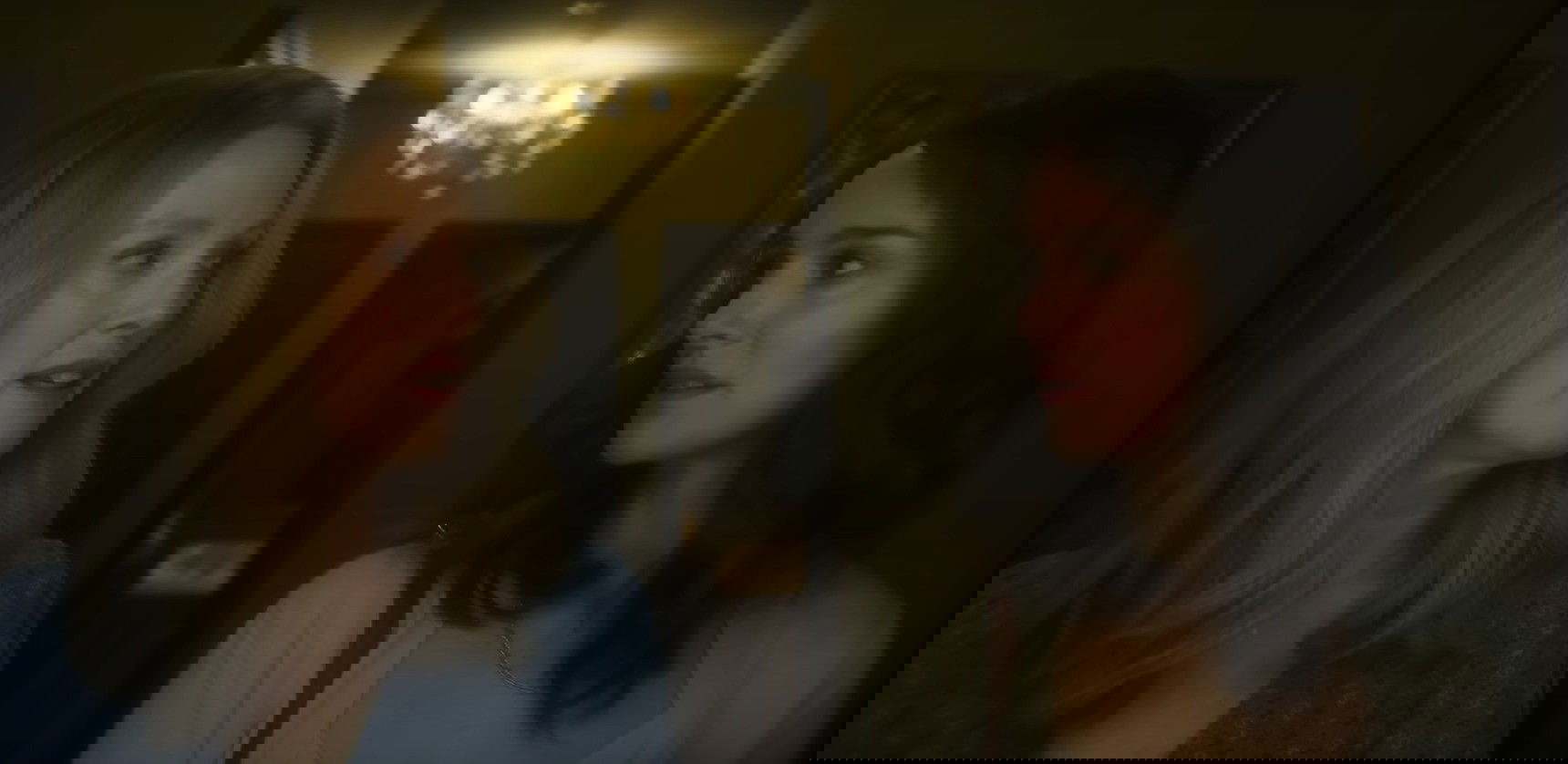 Duas mulheres, uma loira e outra morena, se olham em um ambiente escuro com um lustre ao fundo