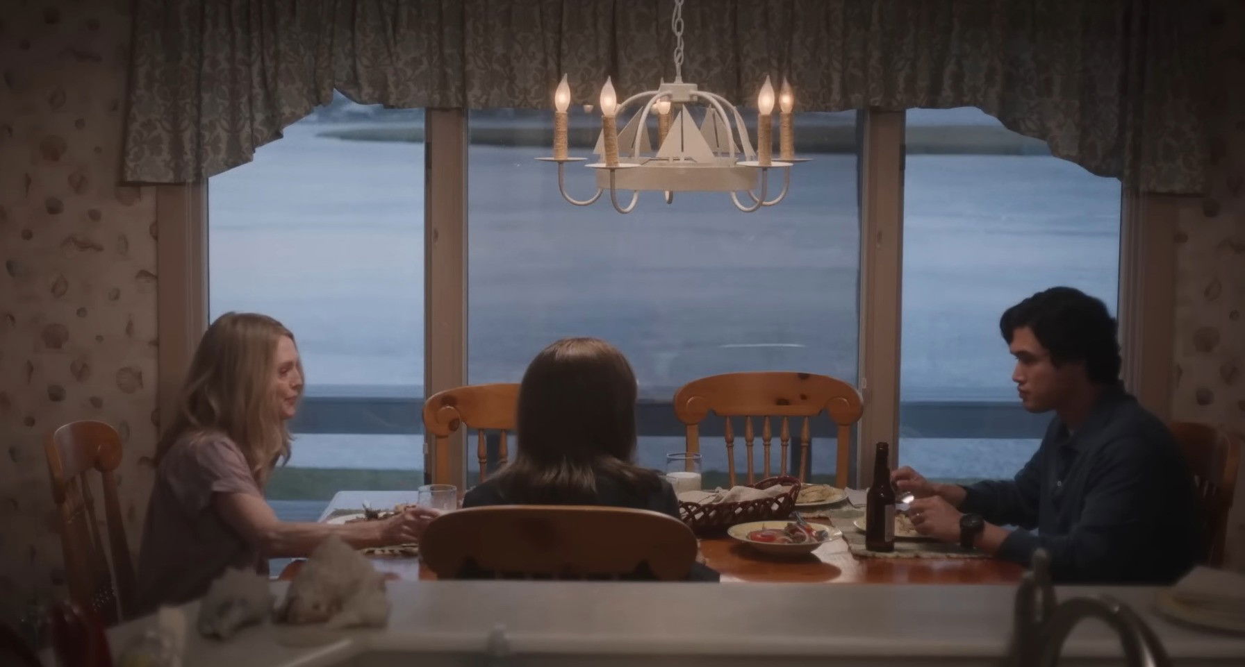 Sala de jantar, com cortinas brancas e  janela ao fundo, e tres pessoas sentadas à mesa de madeira